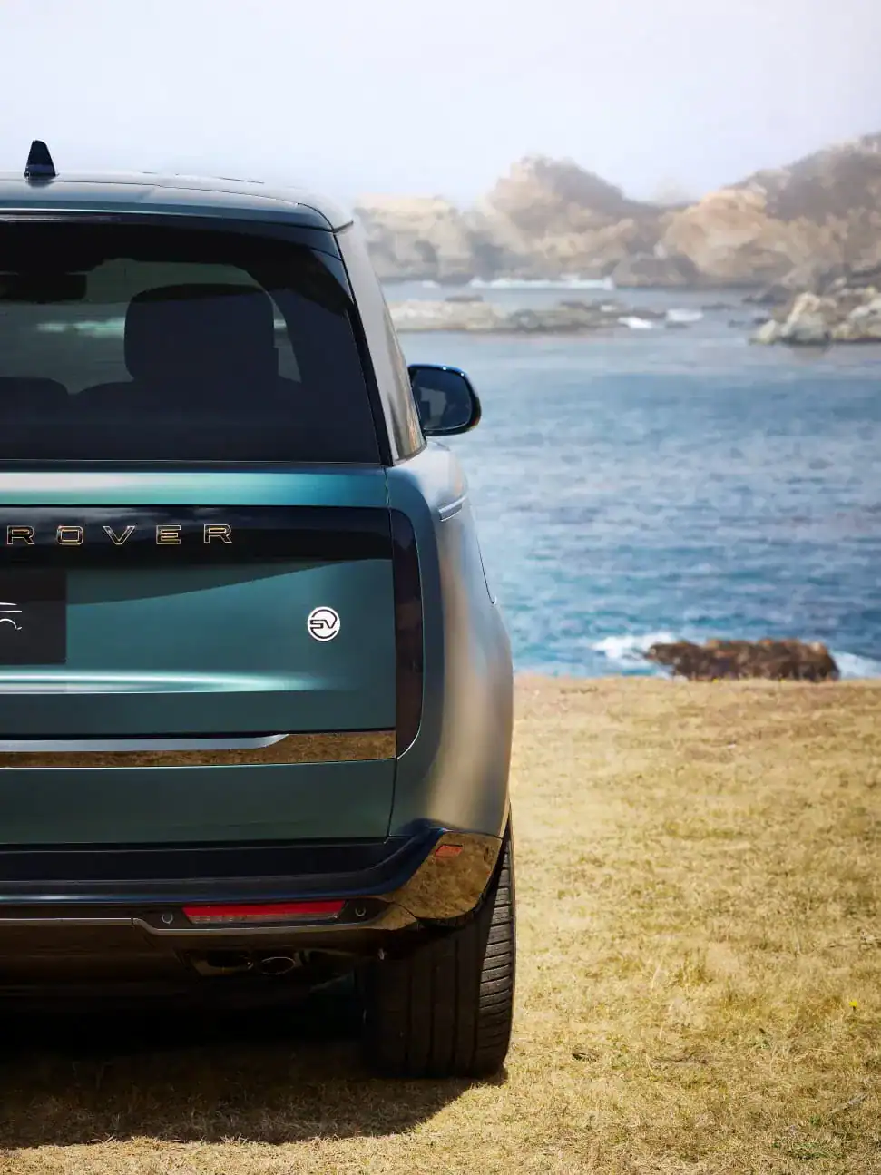 Rear view of a teal luxury SUV parked on a grassy cliff overlooking a scenic ocean and rocky coastline.